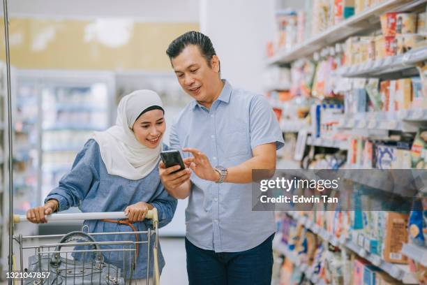 malay couple shopping using smart phone checking shopping list at dairy products section in supermarket during weekend - malay couple stock pictures, royalty-free photos & images