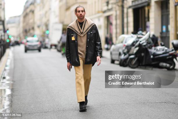 Djebril Irid wears a gold earring, a black wool turtleneck pullover, a gold chain necklace with a moon pendant, a beige ribbed wool V-neck pullover...