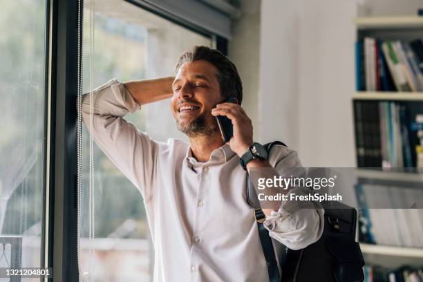 portrait of a handsome young man standing by the window in the library and talking on the phone - relief emotion stock pictures, royalty-free photos & images
