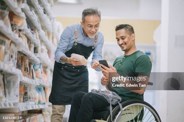 asian indian man with disability on wheelchair asking senior man retail assistant on his shopping list from smart phone at supermarket during weekend - indian shopkeeper stock pictures, royalty-free photos & images