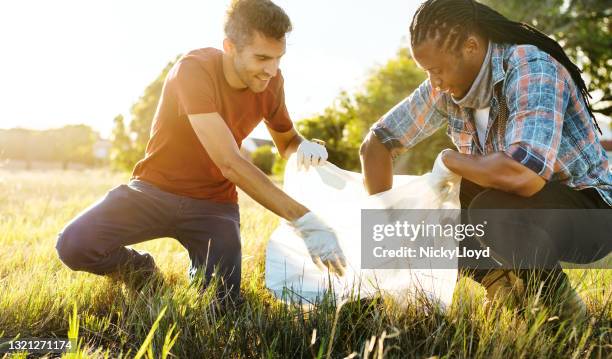 two smiling young male volunteers collecting litter in a field - picking up garbage stock pictures, royalty-free photos & images