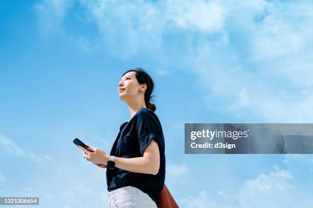 low angle portrait of confident young asian businesswoman using smartphone, looking up to sky with positive mind, standing against blue sky in the city. female leadership and determined to success - looking up ストックフォトと画像