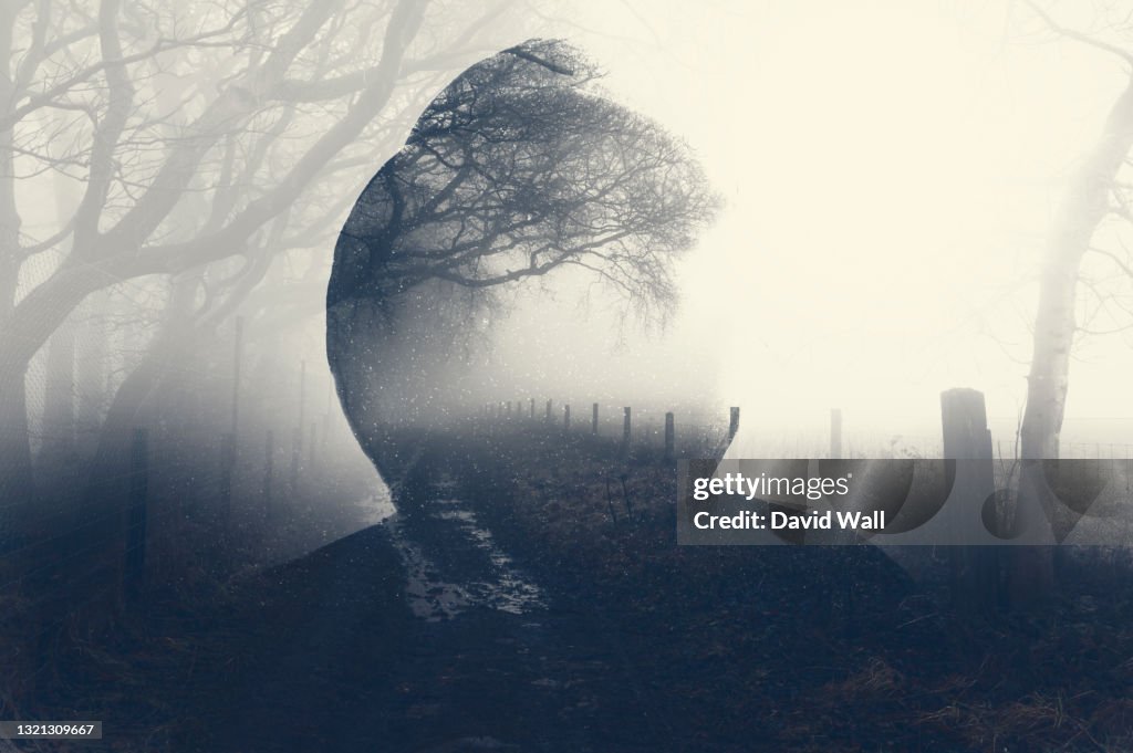 A double exposure of a spooky half transparent hooded figure. Over layered over a foggy path in the countryside. On a moody foggy winters day.