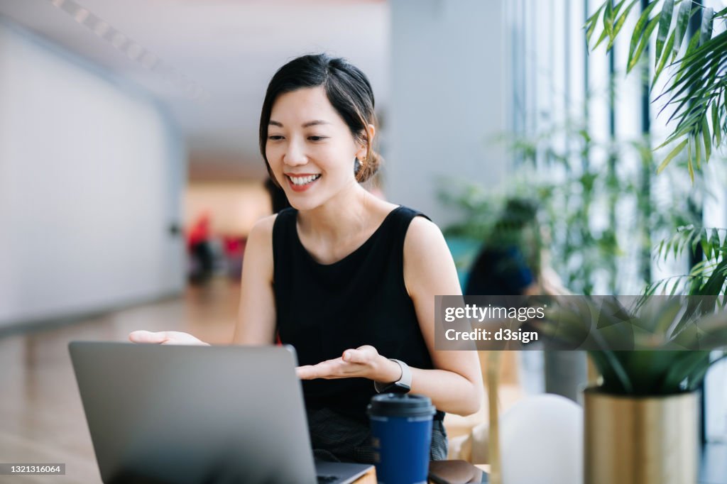 Smiling professional young Asian businesswoman talking in front of the camera having video conference with her business partners on laptop in a contemporary office space