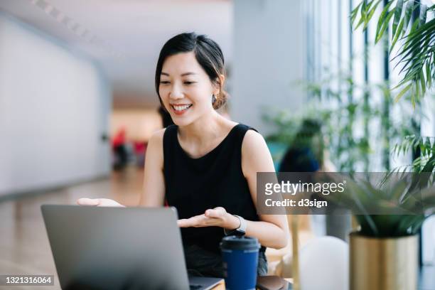 smiling professional young asian businesswoman talking in front of the camera having video conference with her business partners on laptop in a contemporary office space - asia stock-fotos und bilder