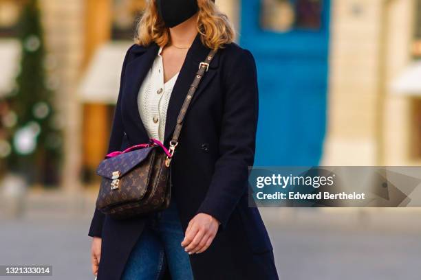 Passerby wears a gold chain necklace, a black face mask, a white wool woven buttoned cardigan, a black long coat, blue denim jeans pants, a brown...