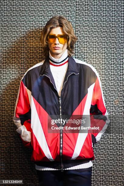 Model wears yellow sunglasses, a white woven wool turtleneck pullover with black and red stripes on the neck, a black red and white pattern oversized...