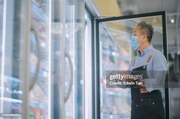 asian chinese senior man with facemask taking inventory with digital tablet at supermarket refrigerated section on frozen food with digital tablet - supermarket fridge stock pictures, royalty-free photos & images