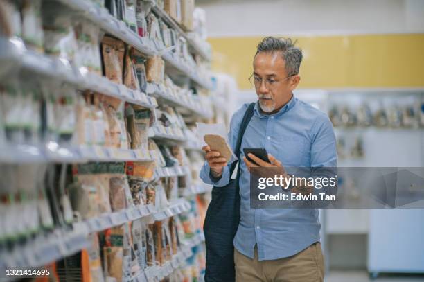 asian chinese senior man checking shopping list using smart phone shopping in supermarket looking for dairy product - comparison shop stock pictures, royalty-free photos & images