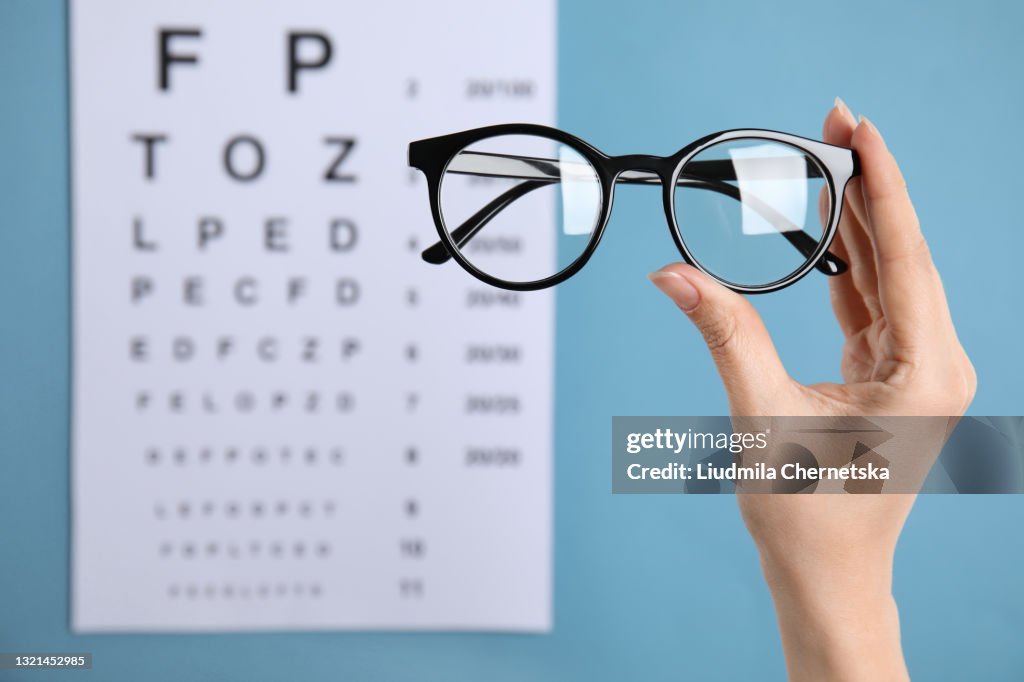 Woman holding glasses against eye chart on blue background, closeup. Ophthalmologist prescription