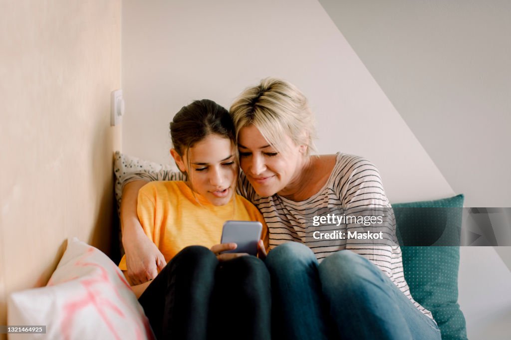 Mature woman sitting with daughter using smart phone in bedroom at home