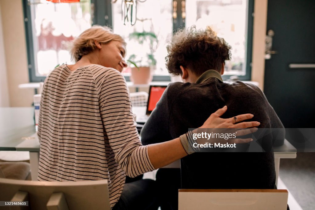 Mother sitting by teenage son studying at home