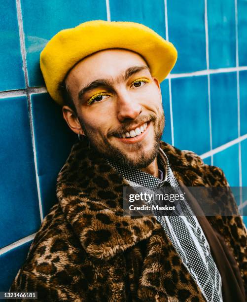 portrait of smiling young man wearing yellow beret against blue tiled wall - beret stock pictures, royalty-free photos & images