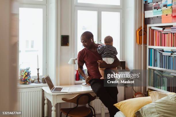 businessman carrying male toddler while telecommuting through laptop at home - pai dono de casa - fotografias e filmes do acervo