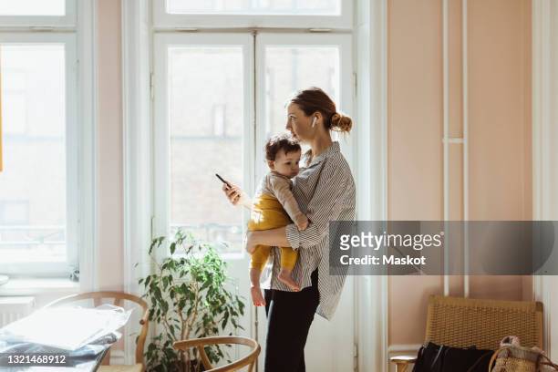 businesswoman carrying baby boy while holding smart phone at home - mãe dona de casa imagens e fotografias de stock