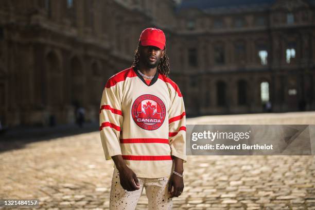 Cali Chiki wears a red Asos cap, a beige with red stripes sport t-shirt with Canada Nations Cup Hockey Tournament logo, white with multicolor paint...