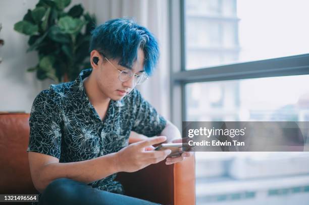 asian chinese teenage boy using smartphone sitting on sofa in living room during weekend - mobile game stock pictures, royalty-free photos & images