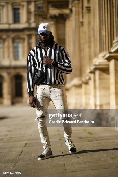 Cali Chiki wears a white and blue denim jean Mitchell & Ness cap, a black and white striped zipped polo shirt, white with multicolor paint spots...