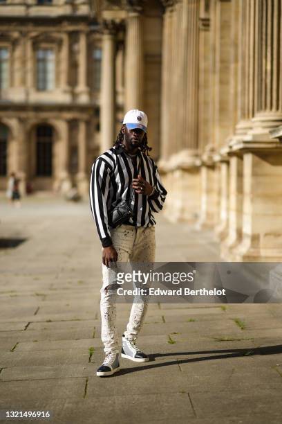Cali Chiki wears a white and blue denim jean Mitchell & Ness cap, a black and white striped zipped polo shirt, white with multicolor paint spots...