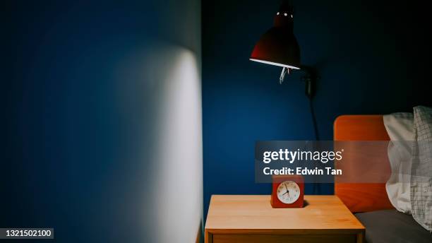 青い壁が付いているベッドの横の夜のテーブルの上の時計と電気ランプによって照らされた夜の寝室 - headboard ストックフォトと画像