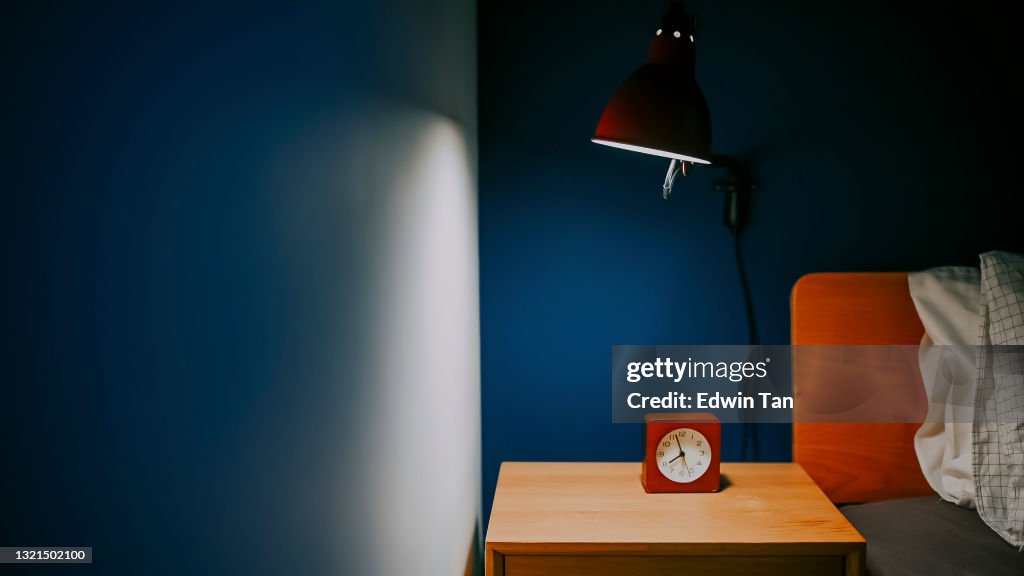 Bedroom at night illuminated by electric lamp with clock on night table beside the bed with blue wall