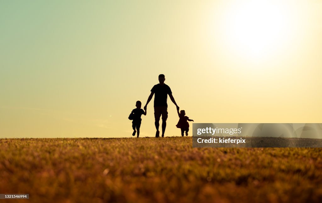 Father walking with son and daughter in park at sunset.