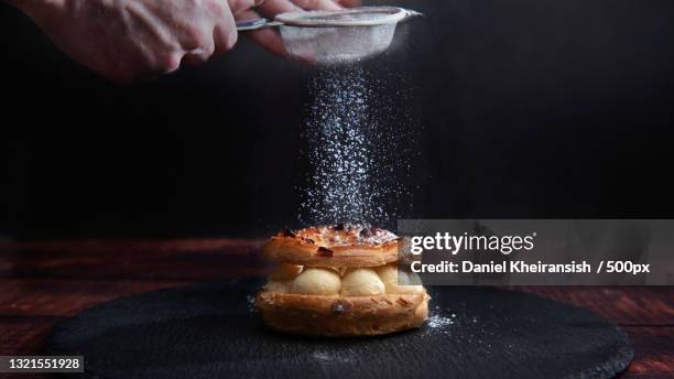 cropped hand of person sprinkling powdered sugar on dessert against black background - azúcar en polvo fotografías e imágenes de stock