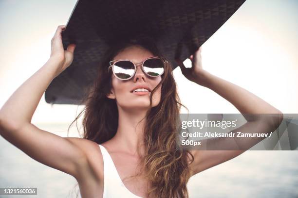 portrait of woman wearing sunglasses and holding surf board above head - sunglasses overhead fotografías e imágenes de stock