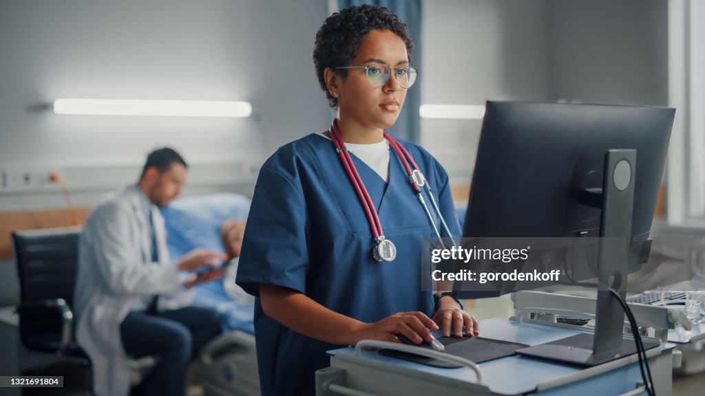 Hospital Ward: Professional Smiling Black Female Head Nurse or Doctor Wearing Stethoscope Uses Medical Computer. In the Background Patients in Beds Recovering Successfully after Sickness and Surgery