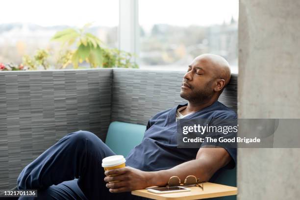 male nurse with disposable cup resting on sofa in hospital - man in hospital stockfoto's en -beelden