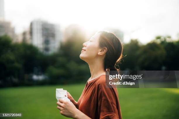 beautiful young asian woman taking off protective face mask, setting herself free and feeling relieved. head up. enjoying some fresh air with her eyes closed while relaxing in park. hope for an end of pandemic concept - end of life care stock pictures, royalty-free photos & images