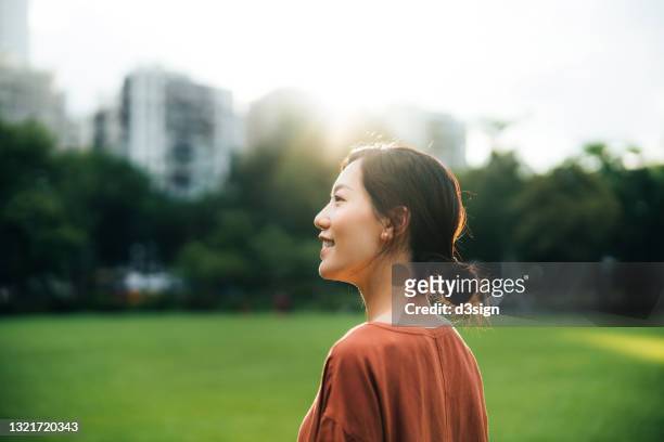 side profile of beautiful smiling young asian woman taking a break in park, looking at view at sunrise, enjoying the beautiful nature. freedom in nature. connection with nature - looking to the future photos et images de collection