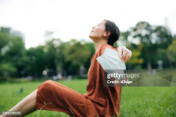 beautiful smiling young asian woman with her eyes closed sitting on the grass in park, taking off protective face mask, setting herself free and feeling relieved. head up. enjoying some fresh air while relaxing in park. hope for an end of pandemic concept - end of life care stock pictures, royalty-free photos & images