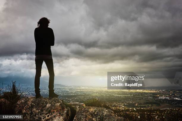 junge frau steht auf einem hohen felsen und schaut auf die sonne, die durch gewitterwolken in der ferne bricht - female looking away from camera serious thinking outside natural stock-fotos und bilder