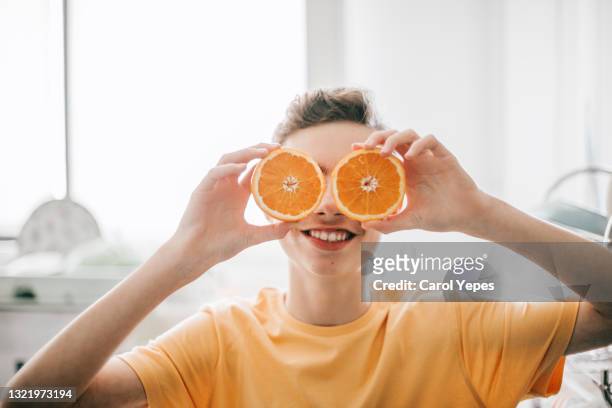 cheerful teenager male holding oranges in front of his eyes at kitchen in the morning - angelica hale fotografías e imágenes de stock
