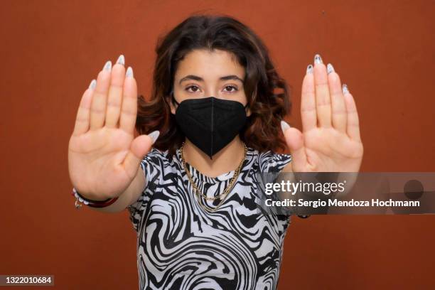 young latino woman wearing a face mask, looking at the camera, and gesturing to stop, red background - hand geste halt stock-fotos und bilder