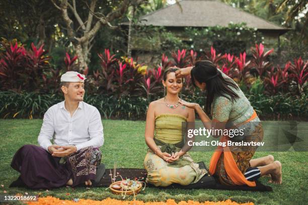 balinese woman in traditional clothing holds blessing wedding ceremony with sacred accessories for european caucasian couple in bali - cerimônia de casamento imagens e fotografias de stock