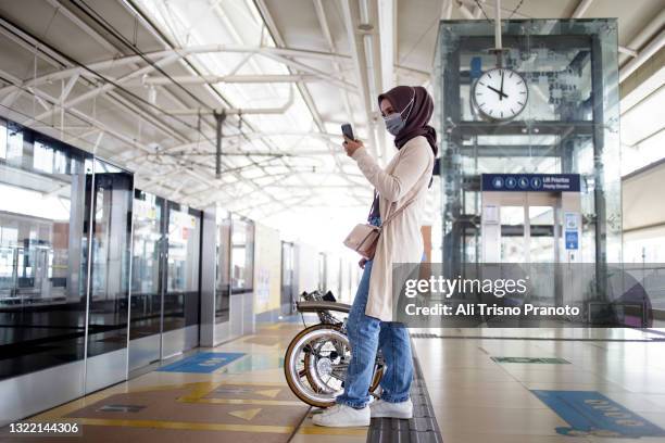 asian young hijab woman, using phone, waiting mrt, with her bike - anständig klädsel bildbanksfoton och bilder