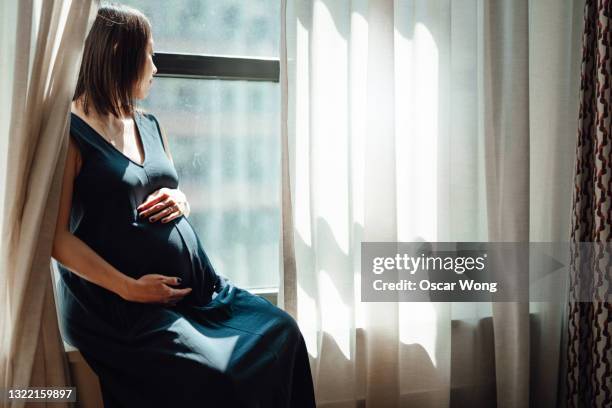 pregnant woman touching her belly while sitting by the window, enjoying beautiful sunlight - woman holding tummy stock-fotos und bilder