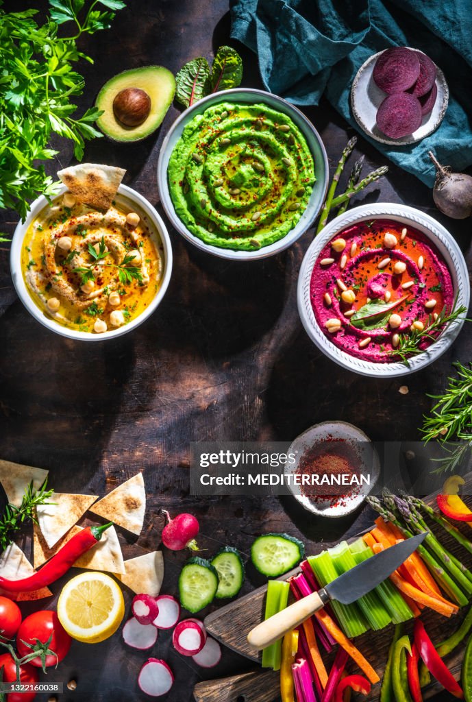 Hummus Three bowl of chickpeas, avocado and beetroot with cut vegetables sticks on dark wood table
