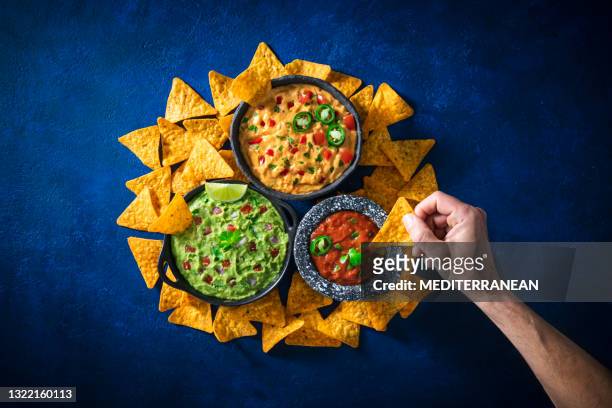 male hand dip in mexican dip sauces guacamole, cheedar dip, tomato salsa with nacho chips - cheese sauce stock pictures, royalty-free photos & images