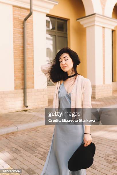 one single stylish young brunette woman in a black hat silk dress and cozy cardigan walks in the middle of empty streets and european buildings enjoying freedom - hair brunette imagens e fotografias de stock