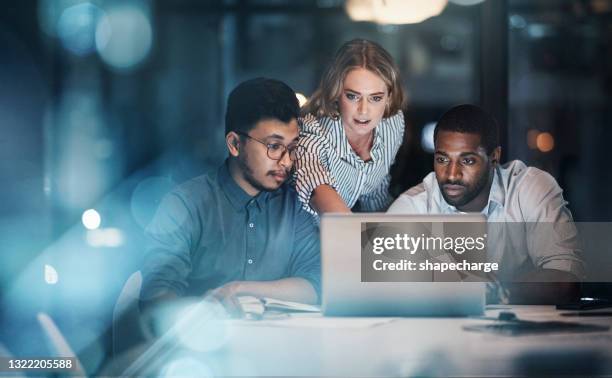 colpo ritagliato di tre giovani imprenditori che lavorano insieme su un laptop nel loro ufficio a tarda notte - cooperazione foto e immagini stock