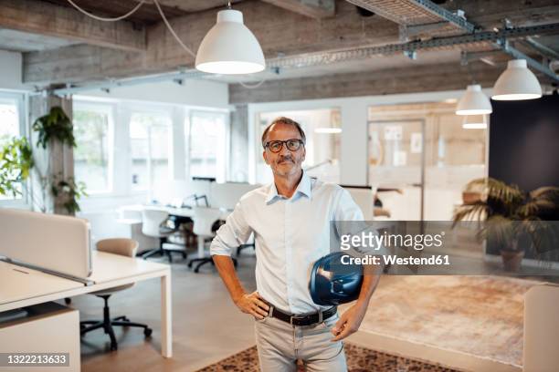 senior male architect holding hardhat while standing with hand on hip at office - architects stock-fotos und bilder