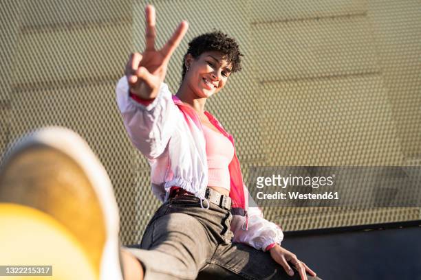 smiling woman showing peace sign while sitting by wall - city wall ストックフォトと画像
