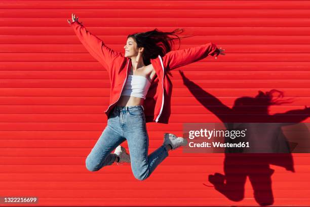 smiling teenage girl with arms outstretched jumping in front of red wall - exhilaration imagens e fotografias de stock