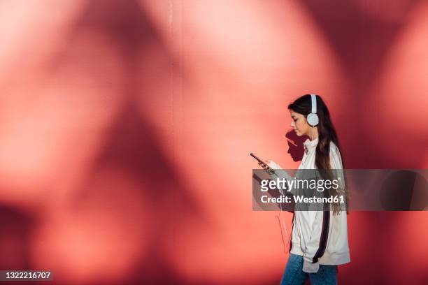teenage girl using mobile phone while walking by red wall - ヘッドフォン ストックフォトと画像