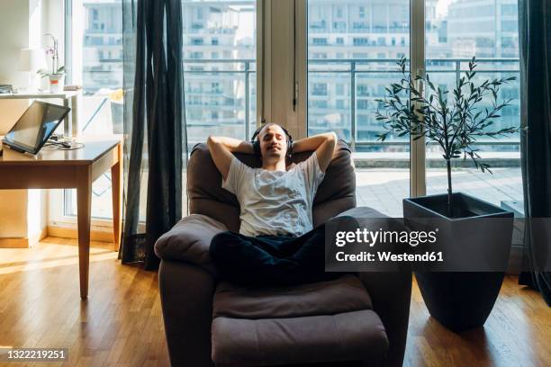 young man with hands behind head relaxing on reclining chair at home - reclining chair stock pictures, royalty-free photos & images