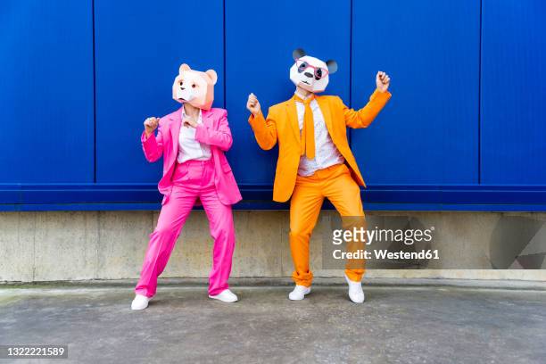 man and woman wearing vibrant suits and bear masks dancing side by side against blue wall - bizzarro foto e immagini stock