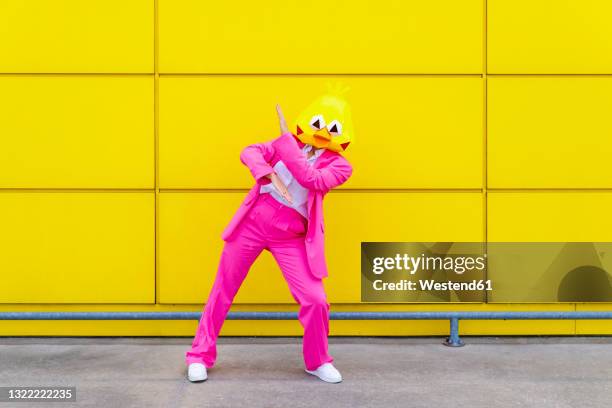 woman wearing vibrant pink suit and bird mask dancing in front of yellow wall - funny mask stock-fotos und bilder